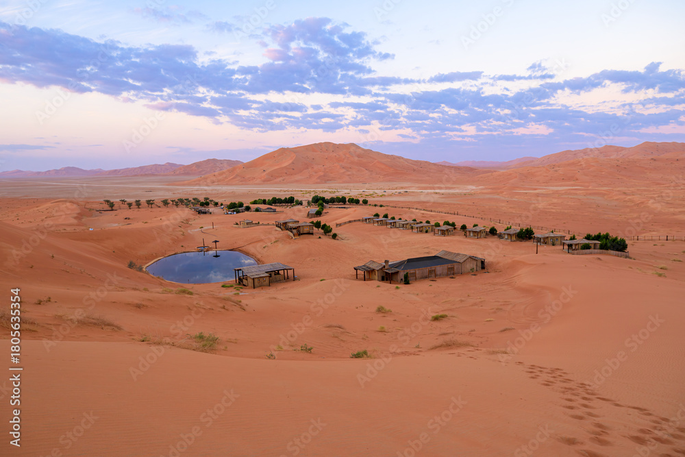 Fototapeta premium Remote desert camp featuring tents and buildings surrounding a refreshing oasis pond, nestled among vast sand dunes in the Empty Quarter of Oman during a tranquil twilight