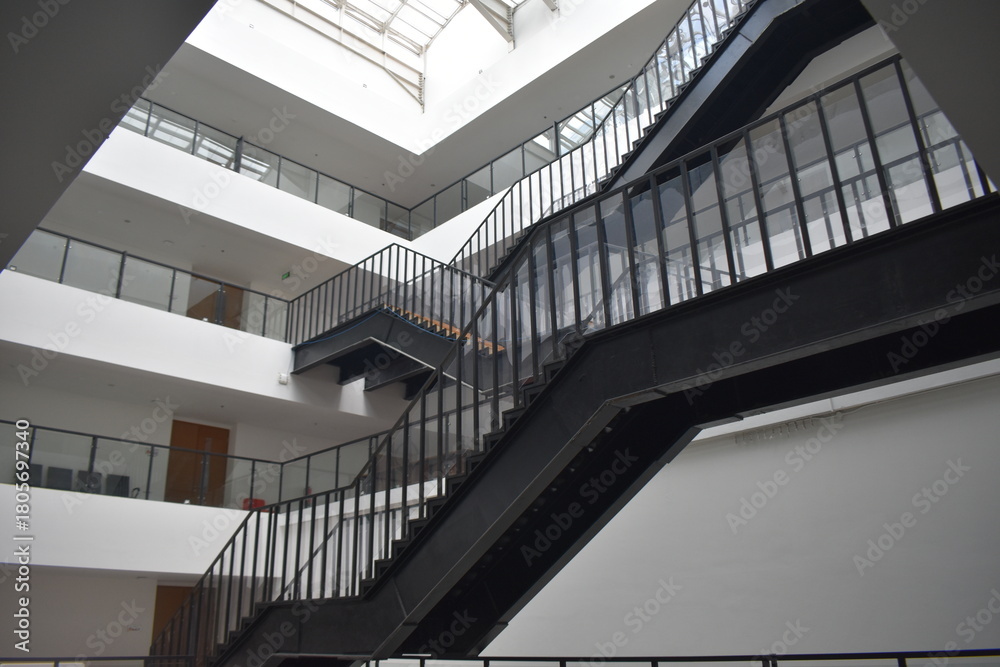 Obraz premium Modern multi-level atrium with sleek black metal staircases, glass railings, and wooden steps. Bright, open space with red seating and minimalist design.