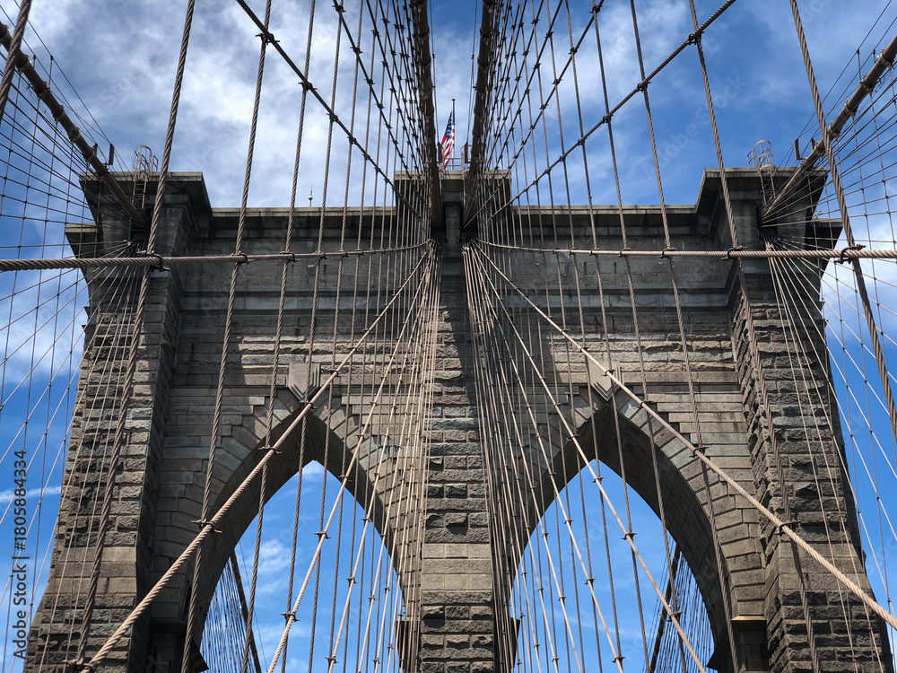 Fototapeta premium An abstract close-up view of a tower and the cables on the Brooklyn Bridge, looking up towards the American flag on a blue sky day