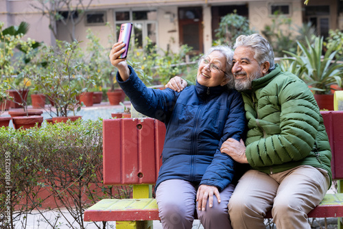 Happy indian senior couple sitting together outdoor in winter season taking selfie picture with smart phone wearing wearing warm clothes. Retirement lifestyle. Active elderly husband wife bonding.