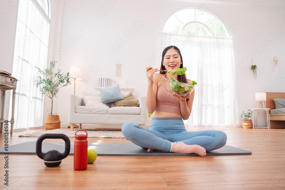 Fototapeta premium Healthy life concept with Asian woman in fitness on yoga mat and eating vegetable salad at home. clean living, nutritional balance, and mindful routine.