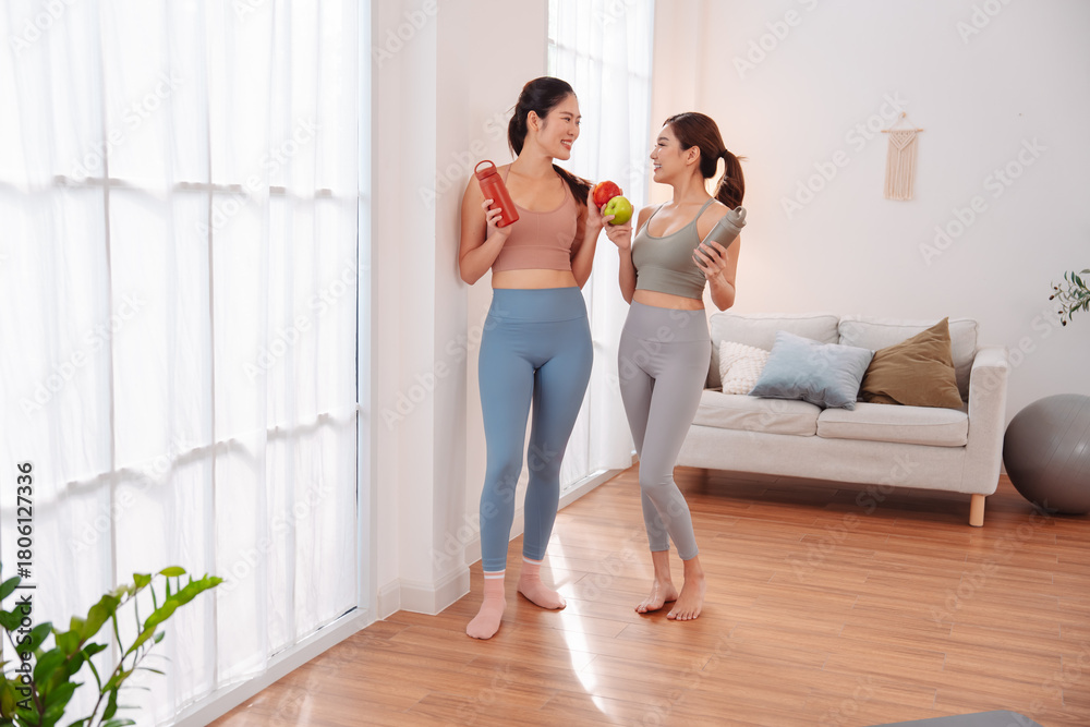 Obraz premium Women talking and laughing after home workout, wearing activewear and holding water bottles in cozy living room. Friendship, healthy lifestyle, wellness, and motivation for staying active together