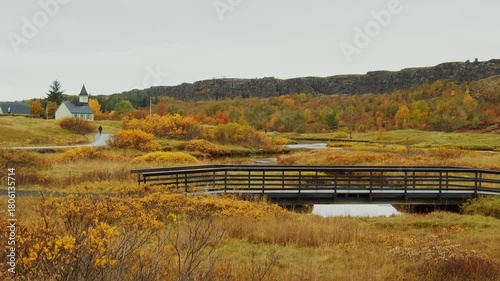 A serene shot of the autumn landscape at Þingvellir National Park in Iceland. The scene features a wooden bridge crossing a small stream, surrounded by vibrant yellow and orange foliage.