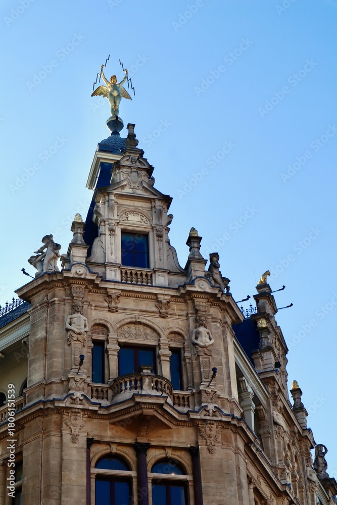 Fototapeta premium Ornate historic building facade featuring a golden angel statue with lightning bolts against a clear blue sky. Antwerp, Belgium