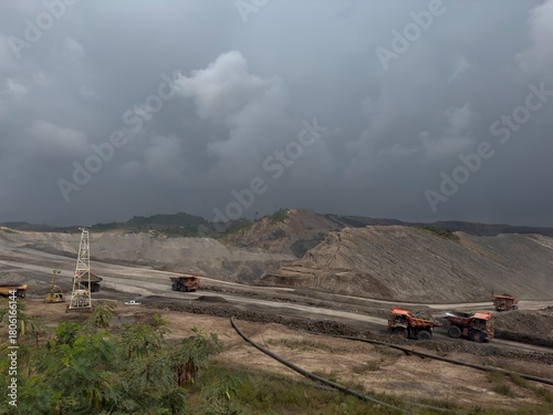 Heavy duty mining trucks operating in a vast open pit coal mine under a dramatic stormy sky.