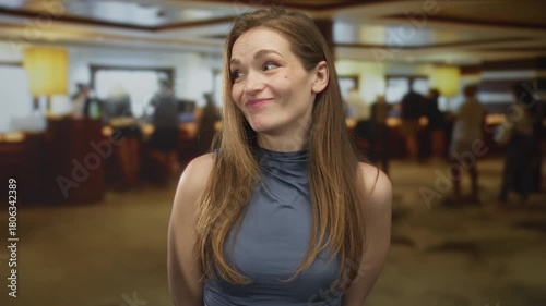 Young caucasian woman makes money gesture by pinching fingertips in hotel building lobby with a skeptical smile; greed.