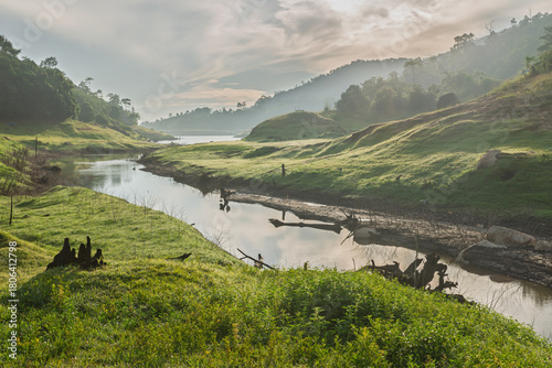 Fototapeta Naklejka Na Ścianę i Meble -  Misty morning sunlight illuminates a lush green valley landscape with a winding stream leading toward distant hills and a reservoir, Serene Nature Landscape.