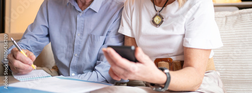 Close up of senior couple managing household finances, researching information on a smartphone, and writing notes