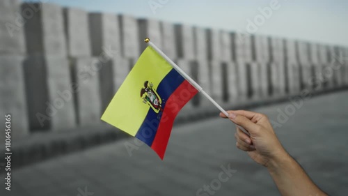 Hand holding ecuadorian flag outdoors near gray monument wall in urban area, symbolizing pride and patriotism in city street environment with male presence.