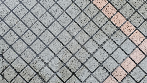 A close-up view of square pavement tiles arranged in a diagonal grid, featuring rough textured surfaces with alternating gray and muted orange tones. The tile floor is wet and damp after the rain.