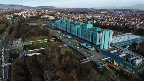 Aerial view of a sprawling, blue-toned hospital complex contrasting against the surrounding autumnal trees and distant cityscape, Zagreb, Croatia.