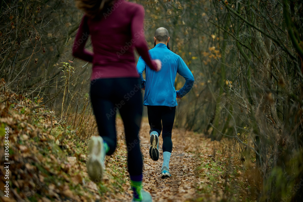 Fototapeta premium Couple running along woodland trail with strong forward stride and focus. Concept of sports and fitness, fall outdoor training, editorial wellness stories and active living.