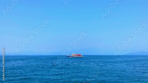 Interesting scene from Lago di Garda with a red, white and blue ferry traversing the lake, leaving wakes between rippling deep blue waters, with impressive mountains visible in the distant background