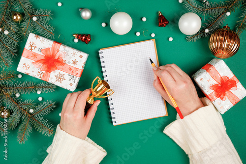 Female hands in white pullover writing in spiral notepad on Christmas green background with copy space. First person top view photo. New years gifts list.