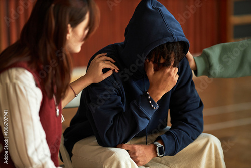 Black teenager boy sitting with head in hand being comforted by Caucasian teenager girl and another teenager, students supporting peer during emotional moment in psychology context