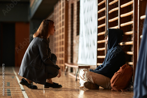 Caucasian young adult woman crouching and talking to seated teenager boy with curly hair and hoodie in gymnasium setting, both engaging in conversation near wooden wall bars