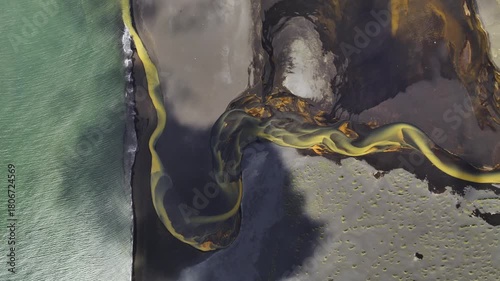 Aerial view of a vibrant yellow river snaking through dark volcanic sands, contrasting sharply with the green ocean waters, Grindavikurbaer, Iceland.