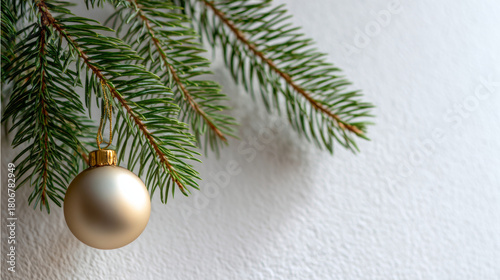 Close-up of green pine tree branches with a single gold Christmas ornament hanging against a textured white background