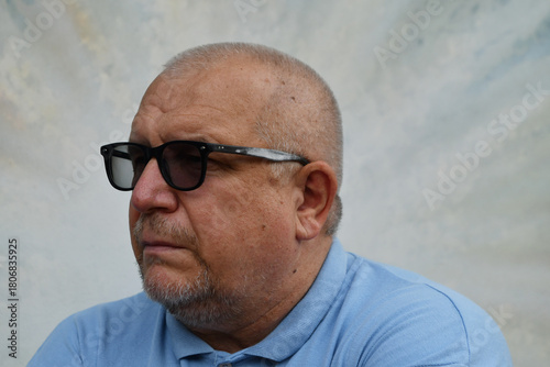 Portrait of a Caucasian elderly man in a blue shirt and glasses on a light background.