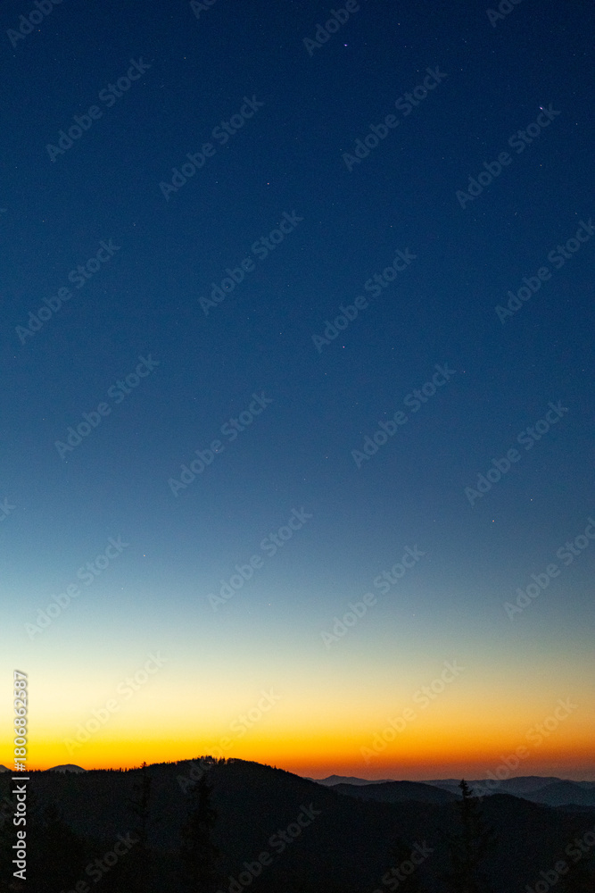 Fototapeta premium Golden band of sunrise edges the horizon beneath a vast blue sky, revealing layered mountain ridges in silhouette. Vertical minimal landscape with faint stars, crisp air, and generous copy space.