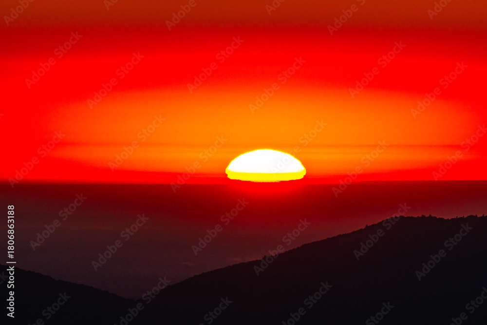 Obraz premium Close view of the glowing sun touching the horizon, in intense red and orange halos above shadowy ridges. Dramatic light and color create an evocative background for travel, wellness, and ads UX.