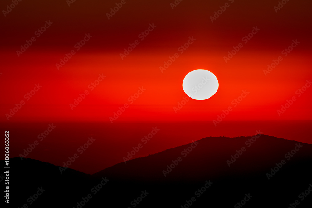 Obraz premium Intense crimson sunset with the sun as a bold white disk above a dark mountain ridge. Minimal, dramatic, and moody, this vivid backdrop suits posters, websites, wellness themes, and travel media.