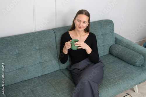 Young woman sitting comfortably on a modern sofa, holding a green mug and enjoying a moment of relaxation at home, taking a break from her daily routine