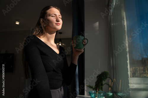 Young woman wearing a black sweater is holding a green mug, standing by a window with a small evergreen tree and enjoying the sunlight and a hot beverage in her apartment