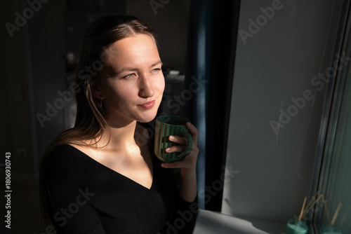 Young woman with long brown hair, wearing a black shirt, holds a green mug and enjoys a quiet moment by the window, basking in the warm morning sunlight streaming into the room