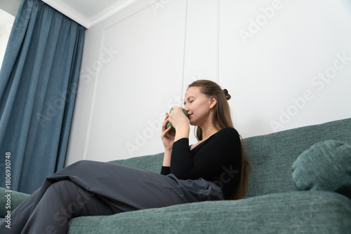 Young woman holding a green mug, savoring the rich aroma of coffee or tea while relaxing on a cozy sofa, cherishing a peaceful moment at home during a quiet afternoon