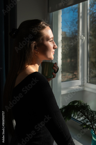 Young woman wearing a black long-sleeved shirt is standing by the window, holding a green mug and looking outside, enjoying the morning sunlight and a hot beverage. Vertical photo