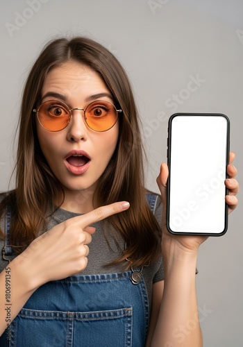 Surprised woman with orange sunglasses pointing at a blank smartphone screen offering a mockup space for advertising or app promotion in a modern presentation
