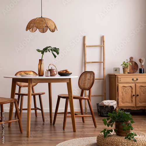 The stylish dining room with round table, rattan chair, wooden commode, poster and kitchen accessories. Beige wall with mock up poster. Home decor. Template.