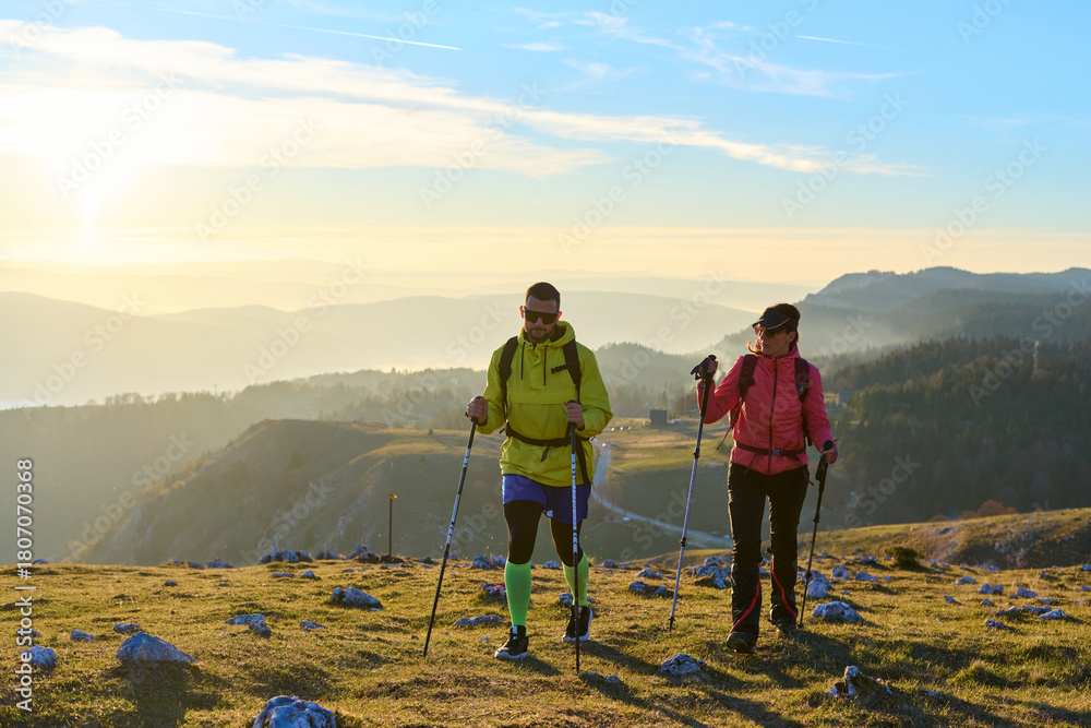 Obraz premium Couple hiking through a grassy mountain trail during sunrise, surrounded by a stunning backdrop of fog in the valleys below. The warm light creates a beautiful atmosphere
