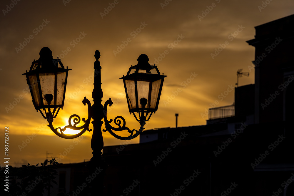 Obraz premium Silhouette of an ornate street lamp against a golden sunset sky in a Mediterranean town. The warm evening light creates a tranquil and nostalgic atmosphere over rooftops and terraces.