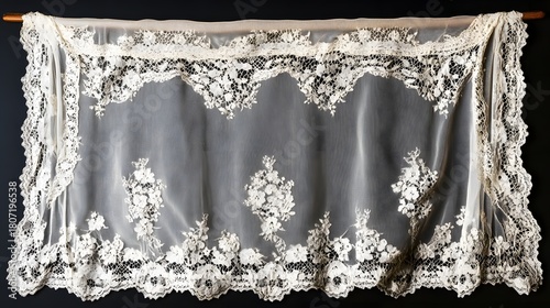 A delicate, wide panel of vintage white lace fabric with intricate floral patterns along the edges, draped and hanging from a wooden rod against a dark background.