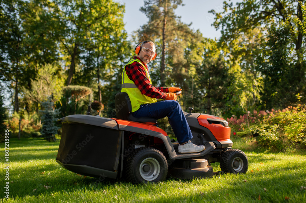 Fototapeta premium Worker in safety gear standing near lawn tractor in garden area