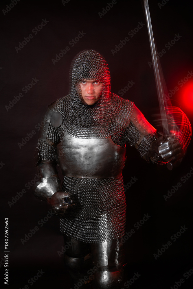 Fototapeta premium A medieval knight in full armor poses against a dark background with red backlighting. Long-exposure photo with motion blur.