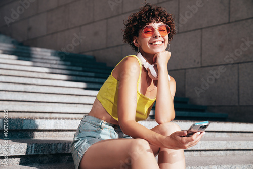 Young beautiful smiling hipster woman in trendy summer clothes. Carefree woman with curls hairstyle, in the street. Positive model holds mobile phone, looks at cellphone screen, uses smartphone apps