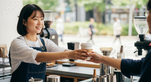 Fototapeta Naklejka Na Ścianę i Meble -  Smiling asian barista handing a takeaway coffee cup to a customer. Friendly woman providing service at an outdoor cafe counter. Small business and coffee culture