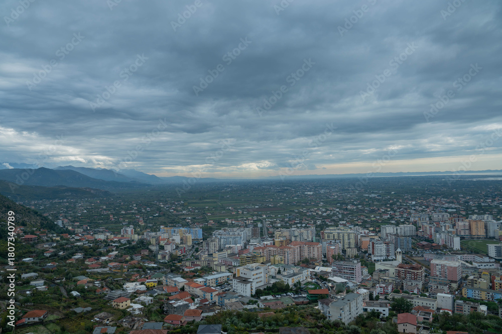 Fototapeta premium A vast, high-angle panoramic shot captures the dense urban area of Lezha, Albania, transitioning into the extensive cultivated coastal plain and valley beyond, all beneath a sweeping, moody sky heavy 