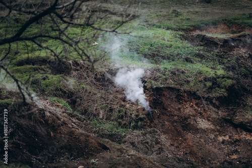A rugged hillside emits white smoke from multiple cracks in the earth, creating a dramatic and surreal atmosphere.