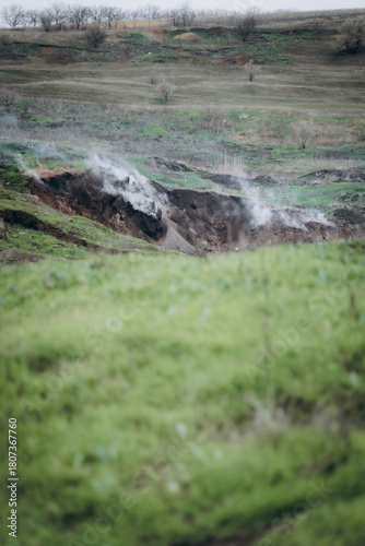 A rugged hillside emits white smoke from multiple cracks in the earth, creating a dramatic and surreal atmosphere.