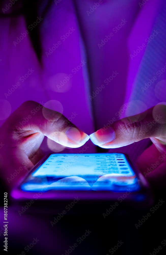 Naklejka premium Close-up of hands typing on smartphone touchscreen at night, glowing blue screen, social media messaging, digital communication, technology lifestyle, modern mobile device, neon purple lighting