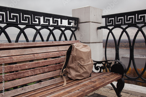 Worn backpack rests on wooden bench in outdoor urban setting. Surrounding pavement is wet, with patches of melting snow nearby. Scene of sense of solitude, mystery, or forgotten moment.