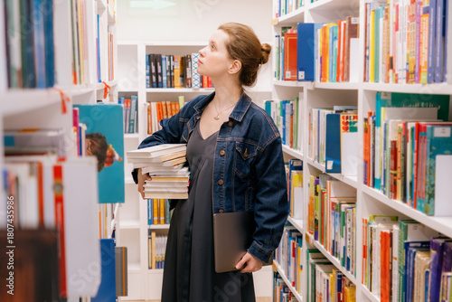 woman holds stack of books among shelves in library. student is getting an education or writing research paper. reading literature. young European female university lecturer.