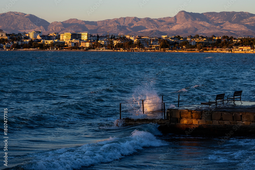 Fototapeta premium Sunlit waves crash against a stone pier at sunset, creating dramatic spray against the dark blue sea, with Side's coastal landscape and mountains glowing in golden evening light. Antalya, Turkey.