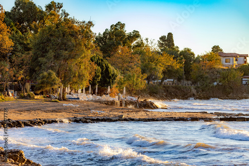 Fototapeta Naklejka Na Ścianę i Meble -  A scenic curved coastline with sandy beach, sunbeds and traditional Mediterranean houses under warm evening light, with white-capped waves on turquoise sea. Side, Antalya, Turkey.

