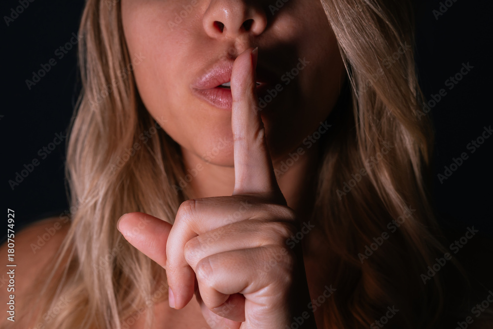 Naklejka premium Close-up of a young woman making a “shh” gesture with her finger on her lips, symbolising silence, secrecy, privacy. Soft lighting, wavy blonde hair, sensual mood on a dark background