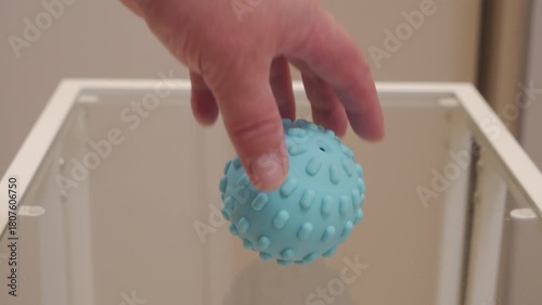 A small, light blue textured rubber or silicone ball is released from a hand and falls onto a clear glass tabletop. The soft, bouncy material makes a very quiet thump as it contacts the hard surface.